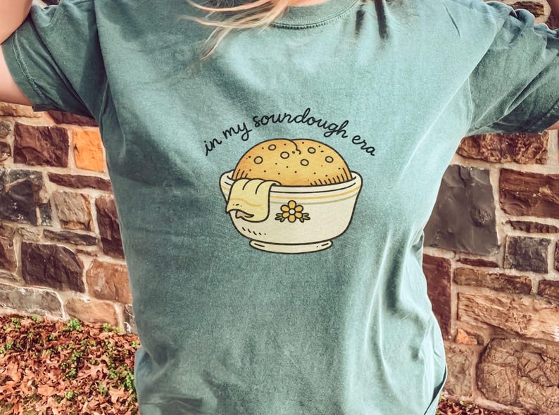 May include: A teal t-shirt with the phrase "in my sourdough era" above a graphic of a sourdough loaf in a bowl. The bowl has a floral design and a cloth draped over the side.