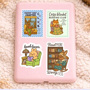 May include: Four colorful stickers with illustrations of animals reading. One sticker says "Rainy Day Chapter Club" with a bunny reading in bed. Another says "Cozy Blanket Reading Club" with a fox reading. The other two say "book lover" and "Read More Worry Less".