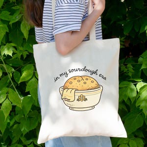 May include: Cream-colored tote bag with the phrase "in my sourdough era" and a cartoon illustration of a sourdough loaf in a bowl. The bowl is light yellow with a floral design. The bag is held in front of a green leafy background.