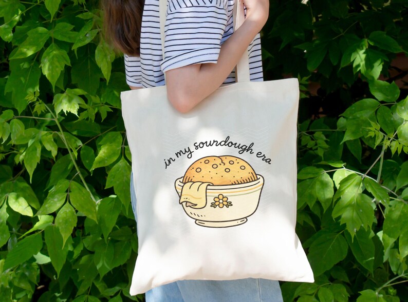 May include: Cream-colored tote bag with the phrase "in my sourdough era" and a cartoon illustration of a sourdough loaf in a bowl. The bowl is light yellow with a floral design. The bag is held in front of a green leafy background.