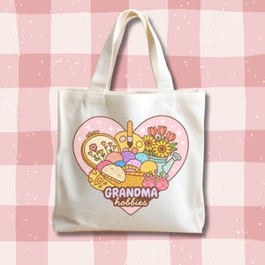 May include: Cream-colored tote bag featuring a heart-shaped design with the words "Grandma hobbies." The design includes illustrations of flowers, yarn, paint, and embroidery, suggesting a crafting theme. The bag has a simple, practical design.