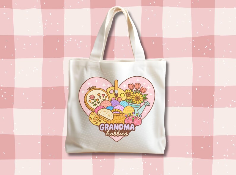 May include: Cream-colored tote bag featuring a heart-shaped design with the words "Grandma hobbies." The design includes illustrations of flowers, yarn, paint, and embroidery, suggesting a crafting theme. The bag has a simple, practical design.