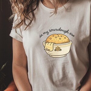 May include: Cream-colored t-shirt featuring a cartoon illustration of a sourdough loaf in a bowl with a towel. The text "in my sourdough era" is arched above the bowl. The design is in a minimalist style.