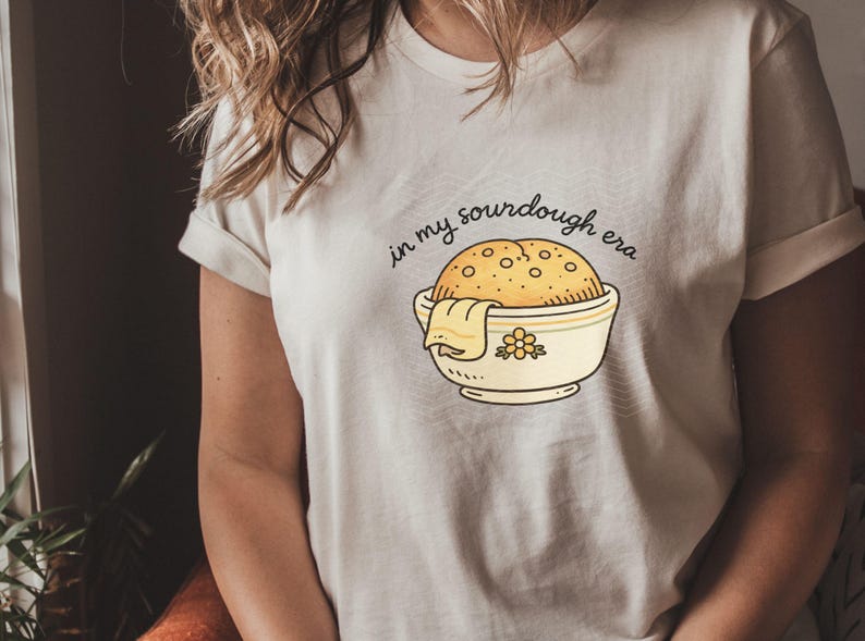 May include: Cream-colored t-shirt featuring a cartoon illustration of a sourdough loaf in a bowl with a towel. The text "in my sourdough era" is arched above the bowl. The design is in a minimalist style.