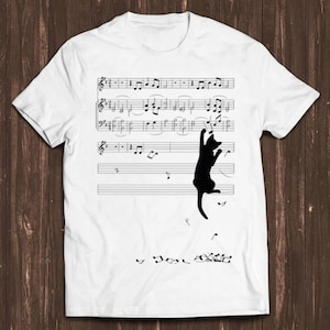 May include: A white short-sleeved t-shirt featuring a black silhouette of a cat reaching for musical notes on a staff. The graphic includes multiple lines of sheet music with scattered notes appearing to fall from the cat's position on the staff.