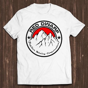 Könnte beinhalten: Ein weißes kurzärmeliges T-Shirt mit einem kreisförmigen Logo auf der Brust. Das Logo zeigt einen roten Hintergrund mit schwarzen Bergsilhouetten, umgeben vom Text RED DWARF und Jupiter Mining Corporation sowie zwei schwarzen Sternen.