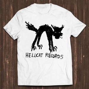May include: A white short-sleeved t-shirt featuring a black graphic of a stylized, arched-back cat with sharp claws and the text HELLCAT RECORDS printed in a hand-drawn, jagged font directly underneath the cat illustration on the front.