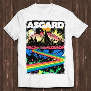 Puede incluir: Camiseta blanca con un diseño gráfico colorido que presenta un camino de arcoíris que conduce a una ciudad con la palabra "ASGARD" en letras negritas.
