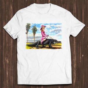 Könnte beinhalten: Ein weißes kurzärmeliges T-Shirt mit einem Grafikdruck einer Person in rosa Kleidung und Hut, die auf einem großen schwarzen Alligator reitet. Der Hintergrund zeigt eine Landschaft mit Palmen unter einem blauen Himmel mit Wolken.