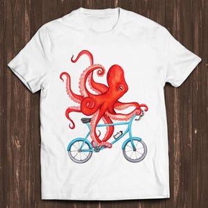 May include: A white t-shirt with a red octopus riding a blue bicycle. The octopus has eight tentacles and is wearing a blue helmet.