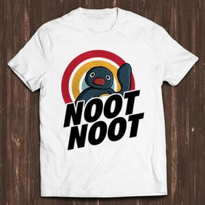 Puede incluir: Camiseta blanca con un pingüino de dibujos animados que lleva un sombrero rojo y amarillo y las palabras "Noot Noot" en negro.