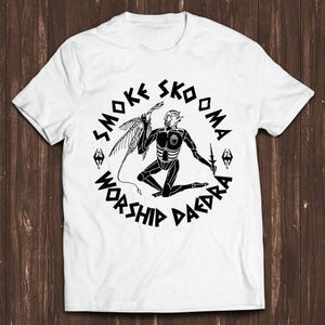 May include: A white short-sleeved t-shirt featuring a black circular graphic. The design depicts a kneeling humanoid figure with a tail and wings, holding a small blade. The text SMOKE SKOOMA WORSHIP DAEDRA is printed in a circular font around the figure.
