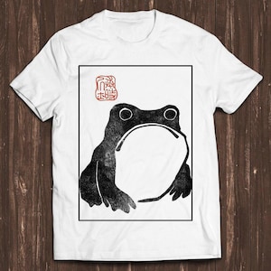 Könnte beinhalten: Ein weißes T-Shirt mit kurzen Ärmeln und einem mittigen, rechteckigen Aufdruck, der eine Frosch-Illustration im schwarzen Tusche-Stil zeigt. Das Design enthält ein rotes, stempelartiges Symbol in der oberen linken Ecke des Rahmens.