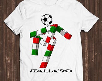 Pode incluir: Uma t-shirt branca de manga curta com um gráfico de um jogador de futebol construído com blocos nas cores verde, branco e vermelho. O jogador tem uma bola de futebol como cabeça e o texto "ITALIA '90" está impresso a preto abaixo.