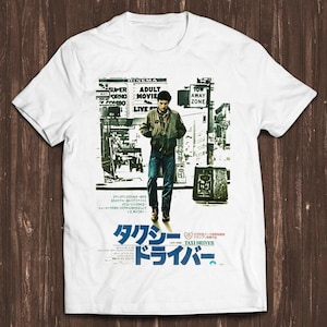 May include: A white short-sleeve t-shirt featuring a black and white graphic print of a person in a jacket and jeans walking down a city street. The print includes text in English and Japanese, including the title Taxi Driver in large blue text.