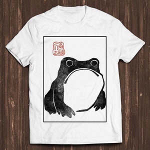 May include: A white t-shirt featuring a black and white image of a frog within a square frame. The image is in the style of a Japanese woodblock print. The text "蛙" is in a red square above the frog.