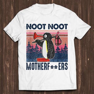 May include: A white t-shirt featuring a graphic of a black and white penguin holding a red trumpet and a plaid bag. The text NOOT NOOT MOTHERF**ERS is printed in dark blue. The background shows a sunset over a forest in shades of pink and blue.