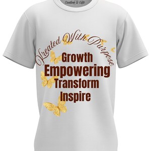 Puede incluir: Camiseta blanca con cuello redondo y mangas cortas. La camiseta presenta el texto "Created With Purpose" en una fuente marrón oscuro curvada, junto con las palabras "Growth, Empowering, Transform, Inspire" apiladas. También hay mariposas doradas con purpurina.