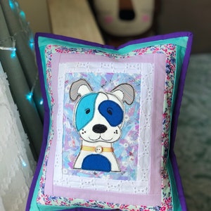 May include: Square decorative pillow featuring a patchwork design with a cartoon dog applique. The pillow has a blue, white, and purple color scheme, with a floral print border and a button detail on the dog's collar. The pillow is approximately 14 inches square.