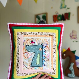 Puede incluir: Un cojín decorativo cuadrado con un diseño de dinosaurio. El cojín presenta un diseño de patchwork con una imagen central de un dinosaurio azul claro con un pajarito. El cojín tiene un borde rojo, verde, amarillo y rosa. El cojín mide unos 35 cm cuadrados.