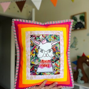 Może przedstawiać: Kwadratowa, kolorowa poduszka z białym królikiem w okularach i koronie. Poduszka ma wzór patchworkowy z warstwami różu, pomarańczy, żółtego i czarno-białą obwódką w paski. Królik ma różową muszkę.