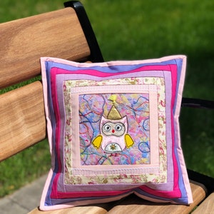 Op de afbeelding: Vierkant decoratief kussen met een geborduurd uilenontwerp. Het kussen heeft een patchwork-ontwerp met roze, paarse en bloemmotiefstoffen. De uil heeft een gouden feesthoed en is gecentreerd op het kussen.