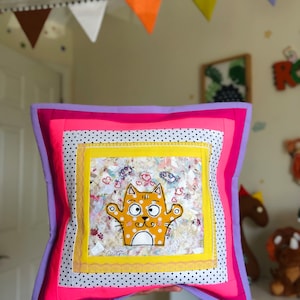 May include: Square decorative pillow with a cartoon cat design. The pillow features a patchwork design with pink, yellow, and purple borders. The cat is orange with white spots and outstretched arms. The background includes hearts and clouds.