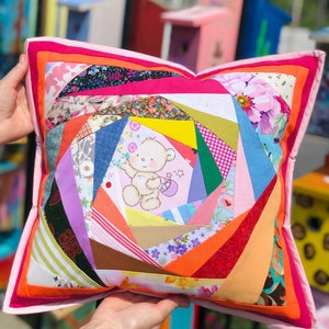 May include: A handmade patchwork pillow with a variety of colorful fabric patterns. The pillow features a central design with a teddy bear illustration, surrounded by geometric shapes in shades of pink, orange, blue, and green. The pillow is held up by a person.