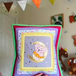 May include: A square, handmade pillow with a whimsical design. The pillow features a central image of a sleeping bear on a gold crescent moon, surrounded by a lavender background and a gold border. The pillow has a green border and is held by a person.