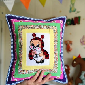 May include: Square decorative pillow with a stitched ladybug design. The pillow features a patchwork design with floral, green, and pink fabrics, and a blue and white striped border. The ladybug is holding flowers.