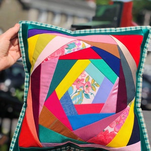 May include: A square decorative pillow with a patchwork design. The pillow features a vibrant array of colors, including red, pink, yellow, blue, and green. The patchwork is framed by a green border with a plaid pattern.