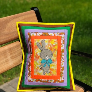 May include: Square decorative pillow with a patchwork design. The pillow features a gray bunny with a blue bow tie on a colorful background. The pillow has a yellow border and is resting on a wooden bench.