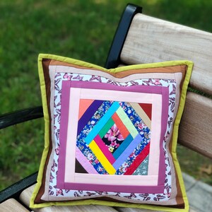 May include: Square decorative pillow with a patchwork design. The center features a colorful geometric pattern with a pink flower. The pillow has a pink, purple, and butterfly patterned border, and a green and brown trim.