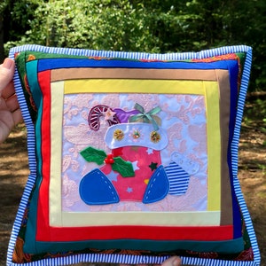 Puede incluir: Un cojín navideño cuadrado y hecho a mano con un diseño de patchwork. El centro presenta un calcetín festivo con acebo, un bastón de caramelo y un lazo. El cojín tiene un borde a rayas y una variedad de telas coloridas.