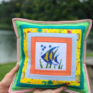 May include: Square decorative pillow with a cross-stitched fish design. The pillow features a white center with a blue and yellow fish, surrounded by orange, blue, and yellow patterned fabric. The edges are green with a striped border.