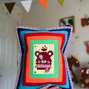 May include: A colourful square cushion with a patchwork design. The cushion features an embroidered bear wearing a scarf, surrounded by orange, green, and blue fabric squares. The border has red, white, and blue stripes. Measures approximately 30 cm x 30 cm.
