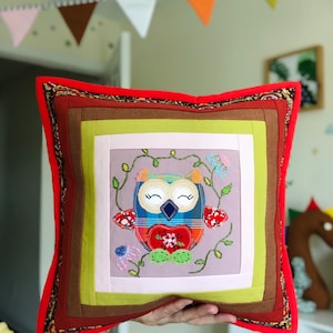 May include: Square decorative pillow with an owl design. The pillow features a patchwork design with a red border and a central image of a whimsical owl with floral and mushroom accents. The owl has a plaid body and a heart-shaped chest.