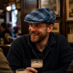 May include: A man wearing a blue plaid flat cap and a navy blue sweater smiles while holding a pint of dark beer. The scene is set in a pub, with framed pictures on the wall in the background.
