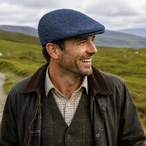 May include: A blue herringbone pattern flat cap. The cap is worn with a dark green jacket with a brown corduroy collar, a plaid shirt, and a gray vest. The background features a green landscape and a cloudy sky.