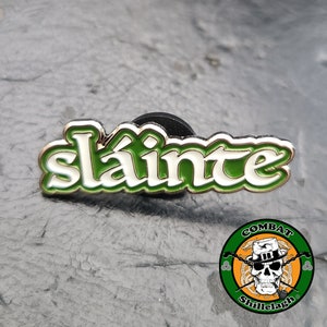 May include: Green and white enamel pin with the word "slainte" in a cursive font. The pin has a gold outline and a black backing.