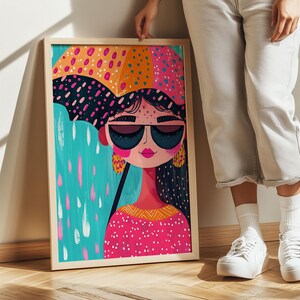 以下が含まれることがあります： カラフルな傘を持った女性が描かれた額装されたイラスト。女性はサングラスをかけ、ピンクと白の模様のドレスを着ています。女性は黒髪で、頬にはピンクのチークが塗られています。