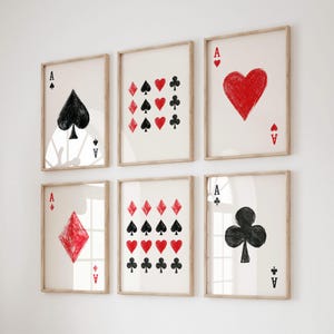 May include: Six framed art prints showcasing playing card designs. Each print features an Ace card with a different suit: spades, hearts, clubs, and diamonds. The artwork is in black and red on a white background, with a simple, hand-drawn style.