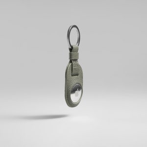 Può includere: Un portachiavi in pelle verde salvia con un portachiavi argentato e un AirTag Apple. La pelle ha una finitura strutturata. L'AirTag è rotondo con un logo Apple argentato. Il portachiavi è progettato per contenere l'AirTag.