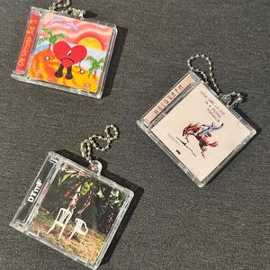 May include: Three clear acrylic CD-shaped keyrings. One features a heart character on a beach scene, another has a horse and rider, and the third shows two chairs in a forest. Each keyring has a silver ball chain.