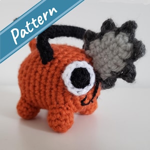 Könnte beinhalten: Ein gehäkeltes Plüschtier in Orange und Grau mit einem schwarzen Griff. Das Spielzeug hat ein großes weißes Auge mit einer schwarzen Pupille und einen kleinen schwarzen Mund.