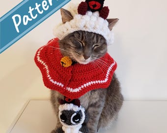 May include: A gray tabby cat wearing a white crocheted hat with a black crocheted bat on top. The cat is also wearing a red crocheted cape with a white trim and a gold bell. The cat is sitting on a white surface and holding a white crocheted sheep with black eyes.