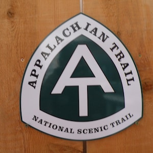 May include: A white and green sign with the text "Appalachian Trail National Scenic Trail" and a white A-shaped symbol on a green background.