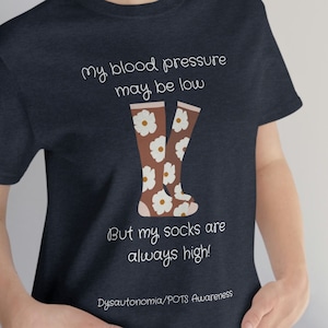Könnte beinhalten: Ein marineblaues T-Shirt mit einer weißen Grafik von einem Paar brauner Socken mit weißen Blumen. Der Text auf dem Shirt lautet "my blood pressure may be low but my socks are always high! Dysautonomia/POTS Awareness".