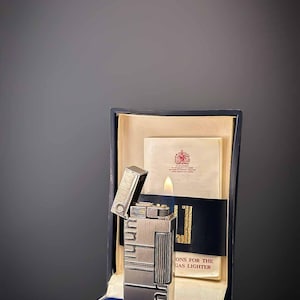 May include: A silver Art Deco lighter with a lit flame, displayed in a navy box with a cream interior. The box includes a booklet and a card with a royal emblem. The lighter has a rectangular shape and geometric patterns.