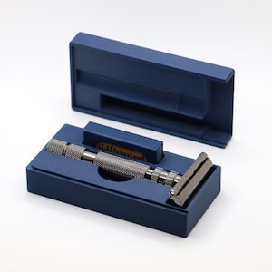 May include: A safety razor with a textured handle and a dark blade, presented in a navy blue box. The box is open, revealing the razor nestled inside a custom-fit compartment. The razor's handle has a knurled design for grip.